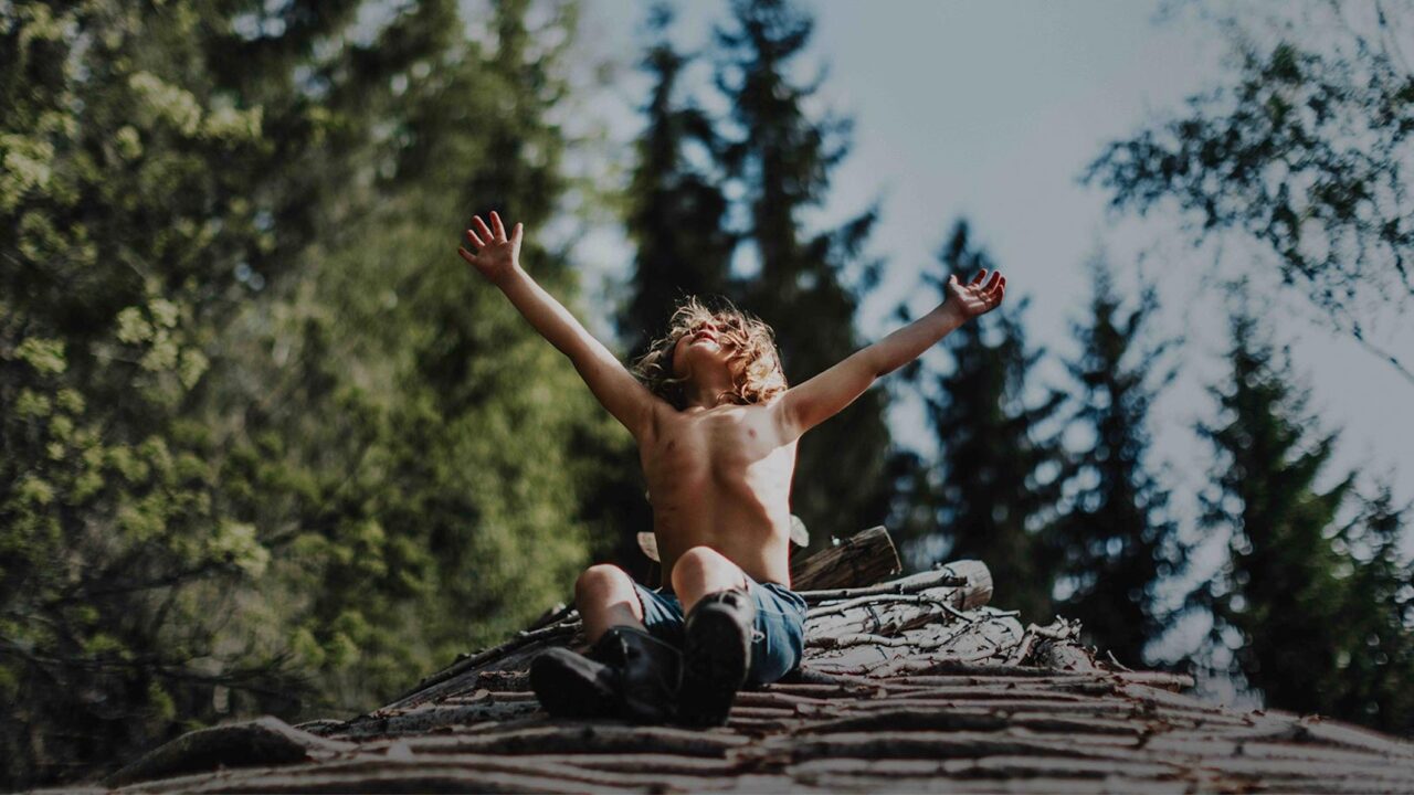 chlapec sedí na kládě stromu s rukama k obloze ve větru